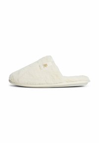 Crème-kleurige fuzzy instapslipper met open achterkant en een klein gouden logo-detail aan de zijkant.