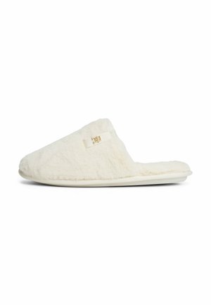 Chausson en peluche de couleur crème à enfiler, avec un dos ouvert et un petit logo doré sur le côté.
