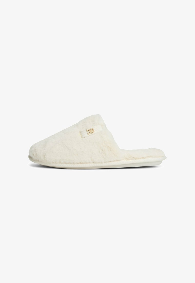 Zapatilla antideslizante de color crema, fuzzy, con parte trasera abierta y pequeño logotipo dorado en el lateral.