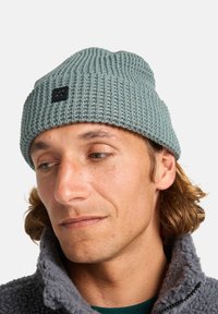 Gorro de punto gris con textura acanalada; presenta una pequeña etiqueta negra con la marca. Material de lana suave, ajuste ceñido. Se lleva con una chaqueta de forro polar.
