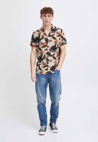 Camisa de boton corto con un fondo beige, que presenta hojas oscuras y flores rojas. Combinada con jeans de color azul claro y zapatillas negras.