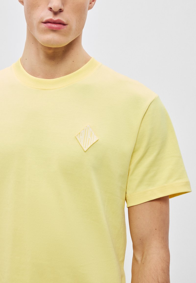T-shirt gialla a maniche corte in morbido cotone. Presenta un piccolo applicazione a forma di diamante sul petto con linee testurizzate.