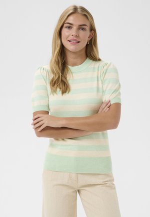 Jeune femme aux longs cheveux blonds portant un pull à manches courtes rayé vert menthe et beige, pantalon beige, les bras croisés, souriante.