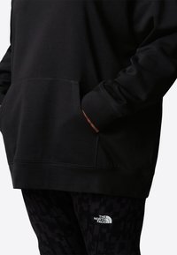 Sweatshirt à capuche noire avec une poche avant, présentant une texture douce et un logo noir assorti. Associé à un pantalon noir à motifs.