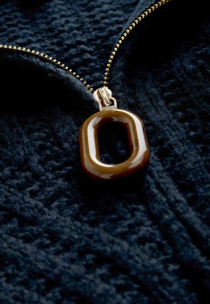 Brauner ovaler Anhänger, der an einem goldenen Reißverschluss auf einem strukturierten, dunkelblauen Stoff befestigt ist und seine glatte Oberfläche sowie das harmonische Farbschema hervorhebt.