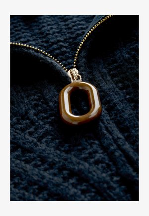 Hnedý oválny prívesok pripevnený na zlatom zipsu na textúrovanom tmavomodrom plátne, zdôrazňujúci jeho hladký povrch a súdržnú farebnú schému.