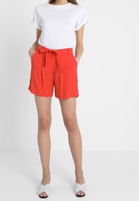 Červené šortky s vysokým pasem, se zavazovacím pásem, skládaným designem, hladkým materiálem a bočními kapsami, v kombinaci s bílým tričkem s ohrnutými rukávy.