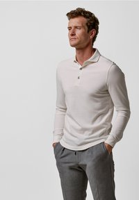 Hellbeige langärmliges Poloshirt mit kragenförmigem Ausschnitt, drei dunklen Knöpfen und einer glatten, weichen Stoffstruktur, kombiniert mit grauen Hosen.