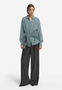 Ljus teal knäppblus med ett subtilt vertikalt mönster, kombinerad med kolgrå vida byxor. En grå stickad tröja knuten i midjan.