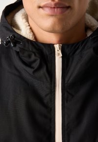 Veste noire fermée avec une fermeture éclair couleur crème et une doublure en polaire portée par une personne, ne montrant que les lèvres et le menton.
