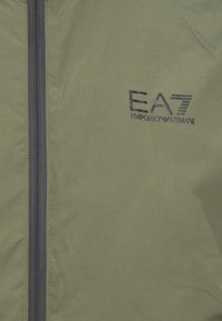 Olivnozelena jakna z gladko teksturo, s središnjim zadrgo in natisnjeno logotipom EA7 Emporio Armani v temno sivi barvi.