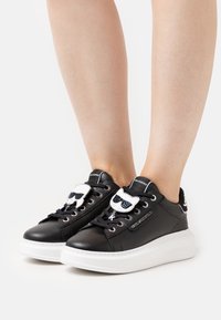Sneakers nere in pelle con suola in gomma bianca, caratterizzate da un emblema a forma di gatto con occhiali da sole sulla linguetta e lacci neri.