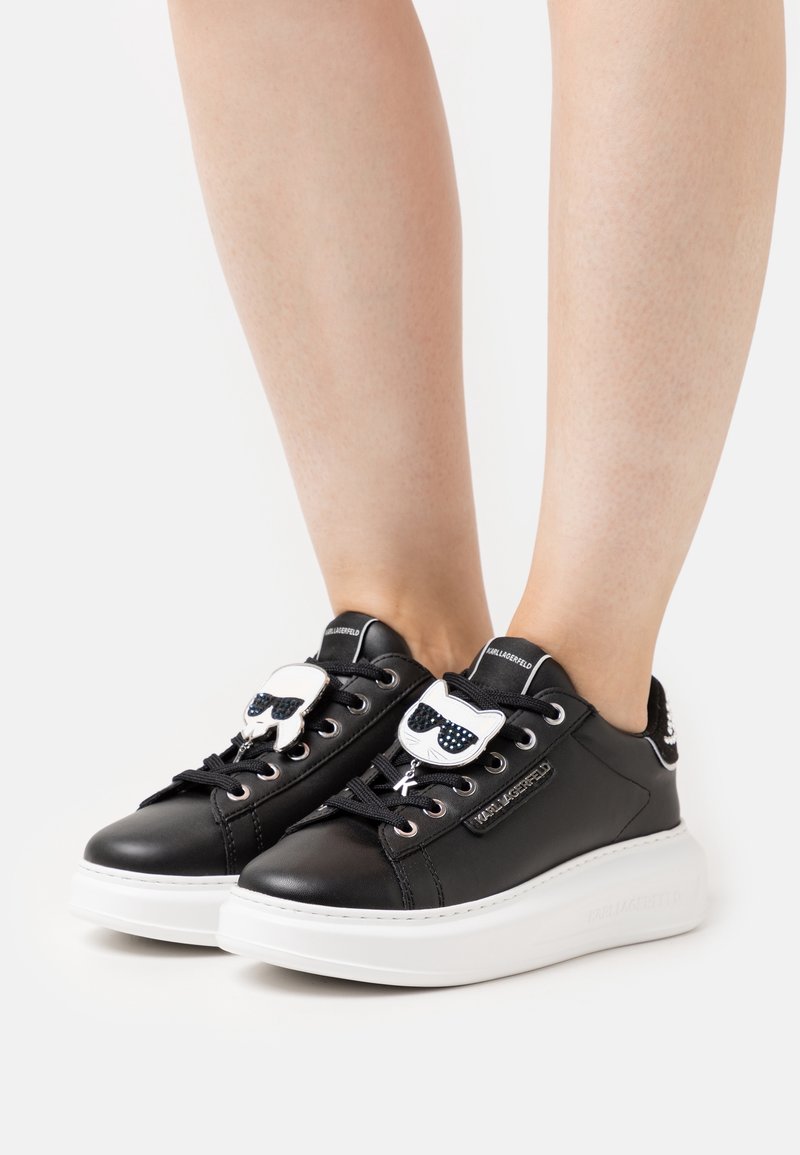 Black leather sneakers with a white rubber sole, featuring a cat-shaped emblem with sunglasses on the tongue and black laces.