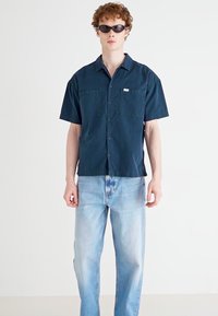 Short-sleeve dark navy shirt with two pockets, button-up front, paired with light blue denim jeans. Model wearing black sunglasses.
