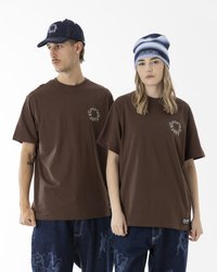 Braune Baumwoll-T-Shirts mit einem runden Logo, kombiniert mit blauen Jeans. Das männliche Modell trägt eine dunkle Mütze; das weibliche Modell hat eine gestreifte Beanie.