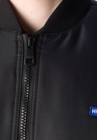 Gros plan sur une veste noire avec un col côtelé, une fermeture éclair visible estampillée "Hugo" et un petit patch logo bleu "Hugo" sur le côté droit.