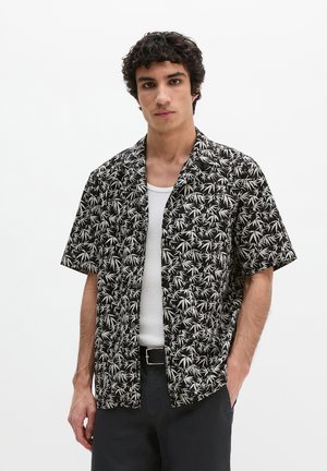 Młody mężczyzna w czarno-białej koszuli w krótkim rękawie z liściastym wzorem, na białym podkoszulku, w czarnych spodniach i z paskiem.