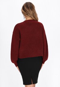 Pulover tricotat de culoare bordeaux cu un design croppat și mâneci tip balon, asociat cu o fustă neagră ce prezintă un detaliu deicătură în spate.