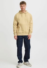 Sudadera beige de algodón con capucha, bolsillo canguro y cordón. Lleva puesta con jeans azul oscuro y zapatillas deportivas de color claro.