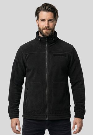 Mann mit Bart, der eine schwarze Fleecejacke mit Reißverschluss und hohem Kragen trägt, steht vor einem einfarbigen grauen Hintergrund.
