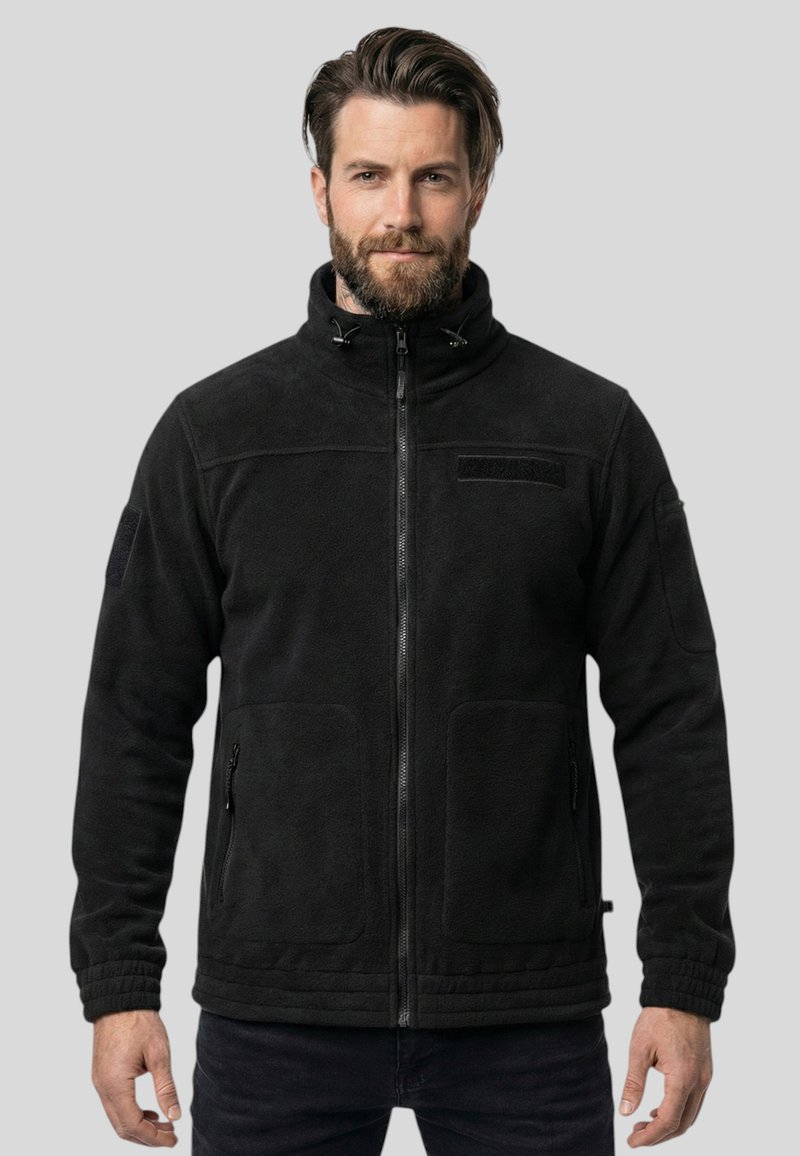 Homme barbu portant une veste polaire noire avec une fermeture éclair et un col montant, debout devant un fond gris uni.