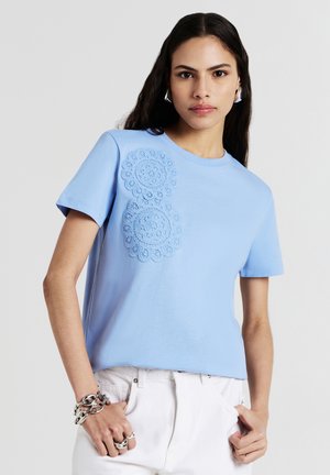 Donna che indossa una camicia azzurra a maniche corte con applique di pizzo floreale e pantaloni bianchi, accessoriate con gioielli d'argento.