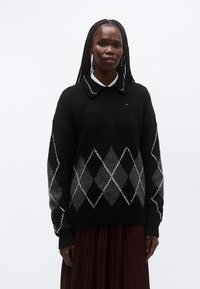 Maglione nero e grigio lavorato a maglia con motivo a rombi, caratterizzato da polsini e bordo inferiore a coste. Indossato sopra una camicia bianca con colletto, per un look a strati.