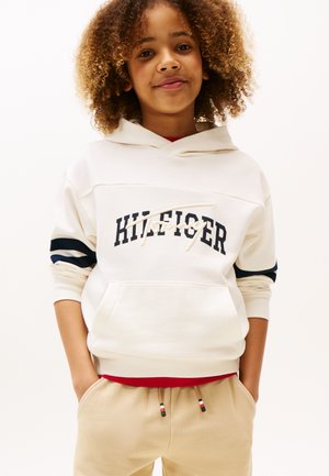 Sudadera blanca con franjas marinas en las mangas, que presenta "HILFIGER" en texto azul oscuro. Lleva puesta sobre una camiseta roja, combinada con pantalones de chándal beige.