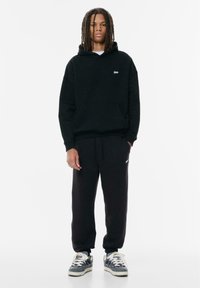 Felpa nera con tasca frontale e piccolo logo, abbinata a pantaloni della tuta neri. Le scarpe sono grigie con accenti crema. Colori solidi, design casual.