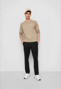 Sweatshirt bege com decote redondo e logotipo, combinado com calças pretas. Finalizado com ténis brancos e um boné bege claro.