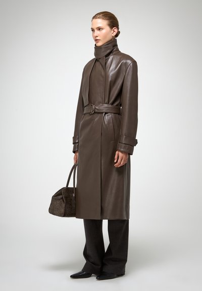 Cappotto trench in pelle marrone con cintura, colletto alto e tasche laterali, abbinato a pantaloni ampi grigi e una borsa in pelle marrone texturizzata.