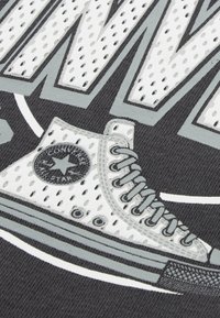 Pelēks grafiskais T-krekls ar stilizētu baltu Converse sneaker ar melniem akcentiem, tumšā fonā ar teksturētiem elementiem.