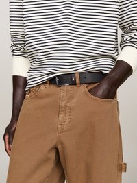 Personne portant un haut rayé à manches longues, un pantalon beige avec une boucle sur le côté et une ceinture noire, avec une main dans la poche et l'autre sur le côté.