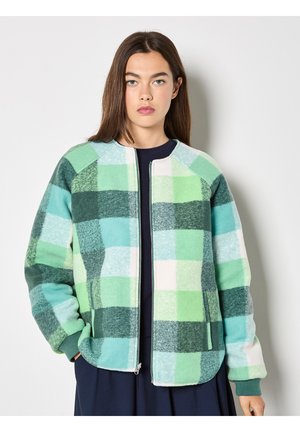 Jeune femme portant une veste zippée à carreaux verts et bleus, les mains dans les poches, debout devant un fond blanc.