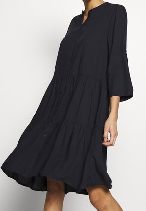 Woman wearing a loose, black, button-up dress with three-quarter sleeves, standing against a plain white background.