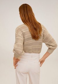 Kobieta z długimi rudymi włosami, ubrana w beżowy sweter o otwartym splocie i białe spodnie z wysokim stanem, stoi z rękami na biodrach, plecami do aparatu.