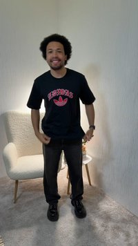 Mann mit Afro-Frisur trägt ein schwarzes Adidas-T-Shirt, schwarze Hose und Schuhe und steht lächelnd neben einem weißen Sessel und einem kleinen weißen Beistelltisch.