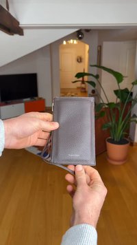 Brunt teksturert lærpung med glidelåslukking, utstyrt med håndleddsrem og preget logo. Vist i en hånd mot et tregulv.