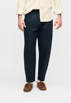 Άνδρας που φοράει μπεζ πουκάμισο με μανίκια γυρισμένα, μπλε-ναυτικό στενό παντελόνι, λευκές κάλτσες και καφέ δερμάτινα παπούτσια για σκάφος, στέκεται μπροστά σε ανοιχτόχρωμο φόντο.