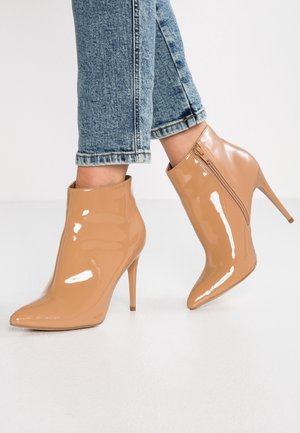 Kvinde iført skinnende nude laklæder ankelstøvler med spidse tæer og høje stiletter, kombineret med blå denim jeans.