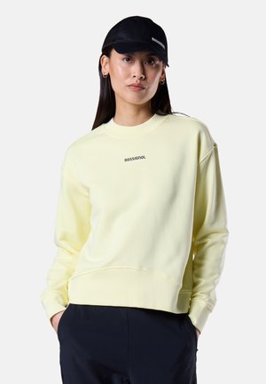 Kvinde iført en lys gul Rossignol-sweatshirt, sort kasket og sorte bukser, stående med hænderne i lommerne mod en ensfarvet baggrund.