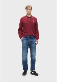 Donkerrood poloshirt met lange mouwen en witte kraagaccenten, gecombineerd met lichtblauwe jeans en zwarte sneakers. Minimalistisch ontwerp, casual fit.