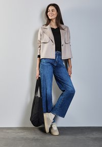 Beige verkürzte Jacke mit Kragen und Fronttaschen, schwarzes Top, weite blaue Jeans, beige Sneakers und eine schwarze Tote-Tasche.