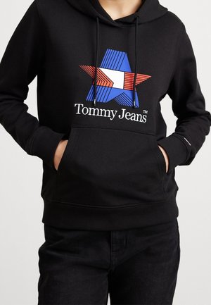 Persona che indossa un cappuccio nero con una stella geometrica blu, bianca e rossa e il testo "Tommy Jeans", con le mani nella tasca anteriore, abbinato a pantaloni neri.