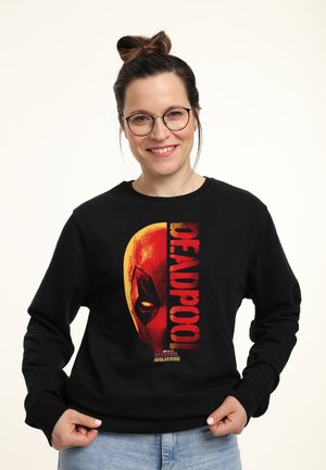 Smilende kvinde med briller iført sort Deadpool-hættetrøje, der viser halvdelen af Deadpools røde maske og tekst.
