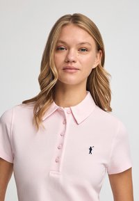 Camisa polo de color rosa claro con cuello, mangas cortas y cinco botones. Presenta un pequeño logo de figura de golfista en azul marino en el lado izquierdo del pecho.