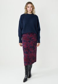 Marineblauer Strickpullover, kombiniert mit einem burgunderfarbenen, floral bedruckten Midirock und schwarzen kniehohen Stiefeln. Der Pullover zeichnet sich durch eine lockere Passform und gerippte Bündchen aus.