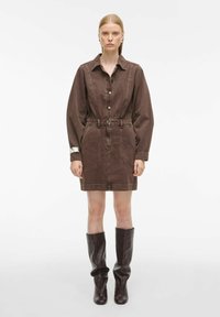 Smeđi traper haljina s dugim rukavima, zatvaranjem na dugmad, pojasom u struku i do koljena. Uparen s visokim smešnim kožnim čizmama.