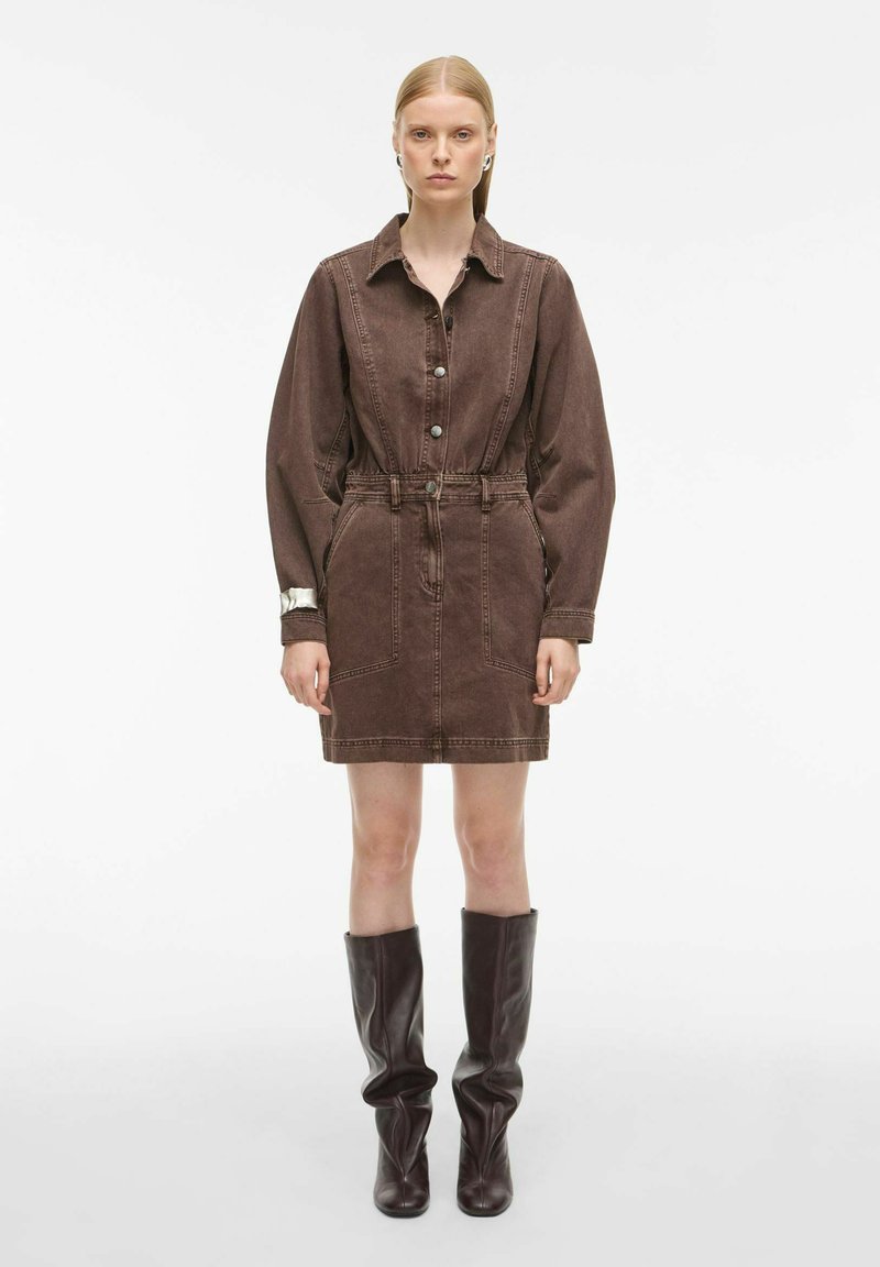 Smeđi traper haljina s dugim rukavima, zatvaranjem na dugmad, pojasom u struku i do koljena. Uparen s visokim smešnim kožnim čizmama.