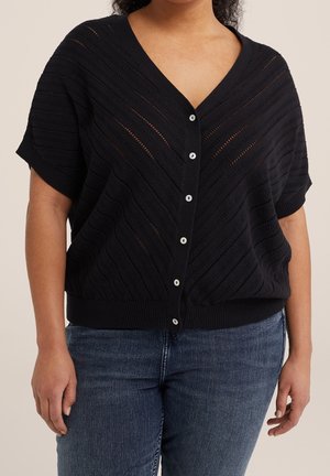 Person trägt eine schwarze, kurzärmelige, gestrickte Strickjacke mit diagonalen Mustern und blauen Jeans, steht vor einem schlichten Hintergrund.